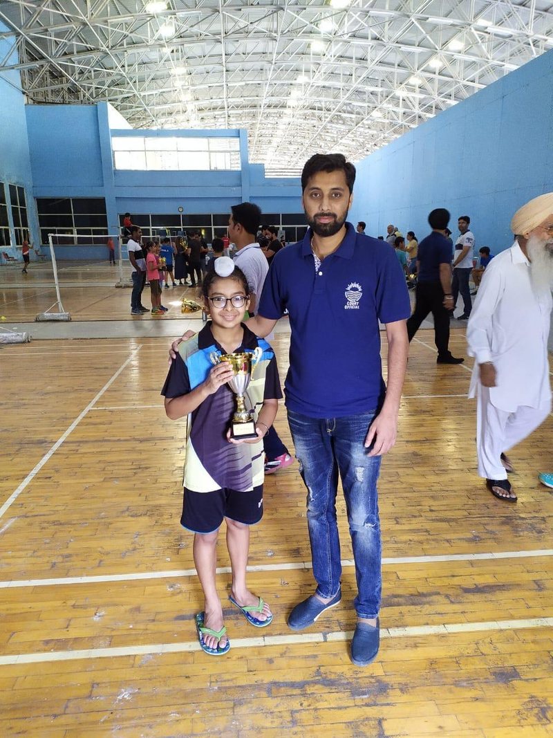 A young Jagsher Singh holding a small trophy beside Coach Suresh Kumar inside a badminton hall